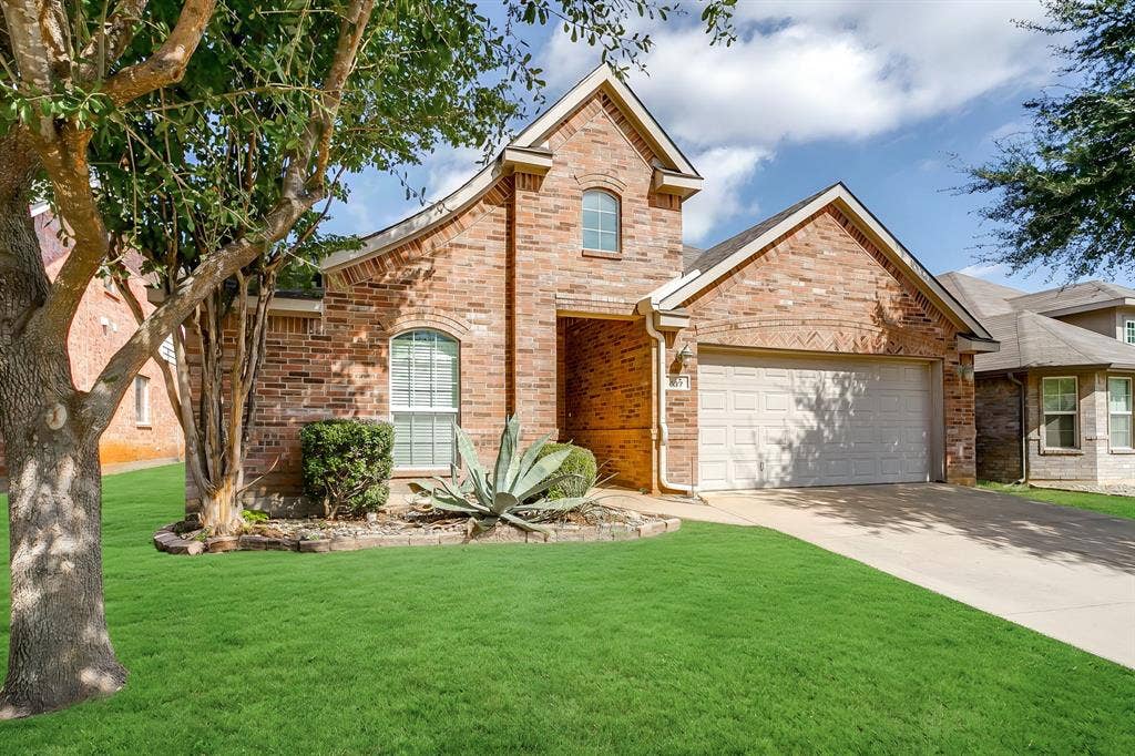 Brick exterior featuring a gabled roofline, arched window detail, concrete driveway, and established landscaping