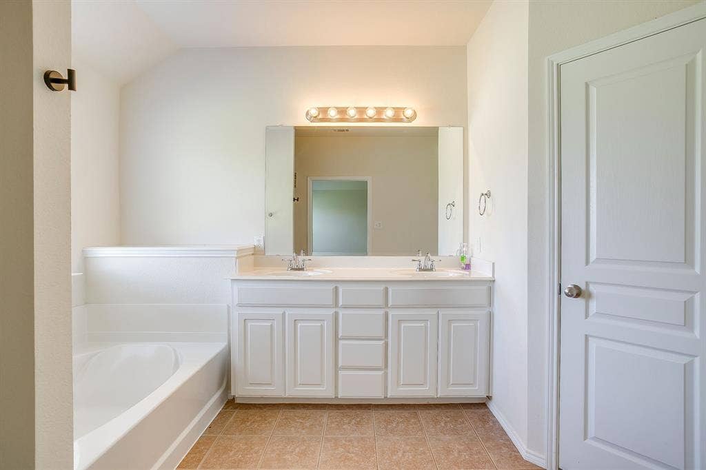 Bathroom featuring a dual-sink vanity with white cabinetry, a large wall-mounted mirror, an integrated soaking tub, and tile flooring