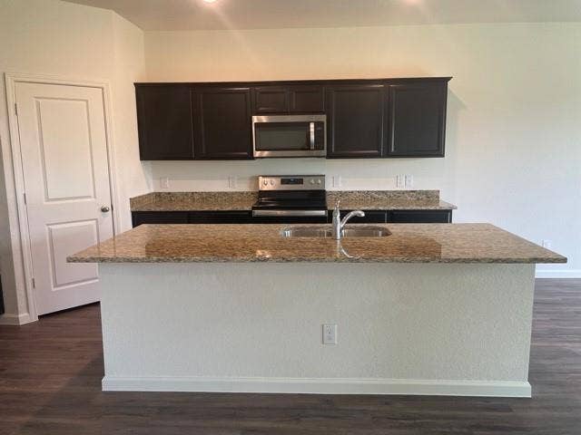 Kitchen featuring dark wood cabinetry, stone-surface countertops, central island with an integrated sink, stainless steel microwave, and wood-finish flooring