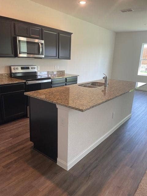Kitchen featuring dark cabinetry, stone-surface countertops, central island with an integrated dual-basin sink, stainless steel microwave, and wood-finish flooring