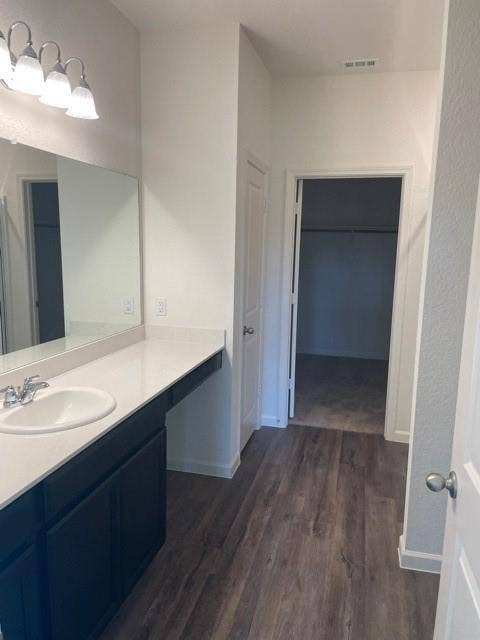 Dark cabinetry vanity with a light-toned countertop, integrated basin, wall-mounted four-light fixture, oversized mirror, and wood-finish flooring