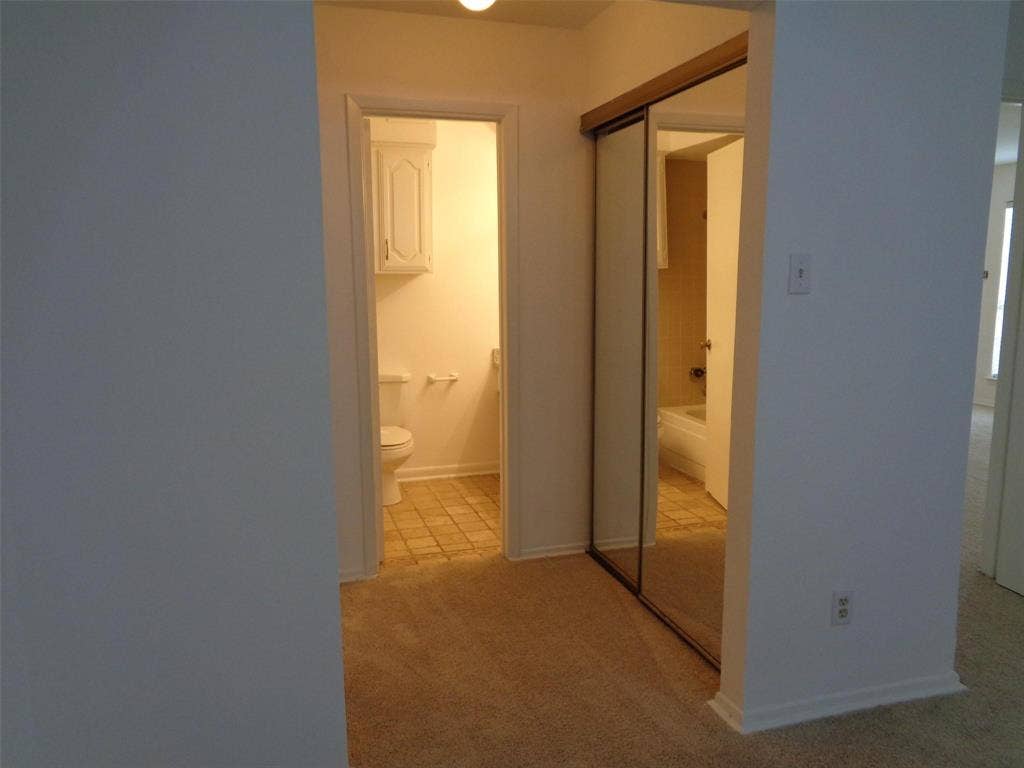 909 Turtle Cove, #141, Irving, TX 75060 Hallway featuring neutral carpeting, floor-to-ceiling sliding mirror closet doors, and direct entry into a tiled restroom equipped with a wall-mounted cabinet and commode