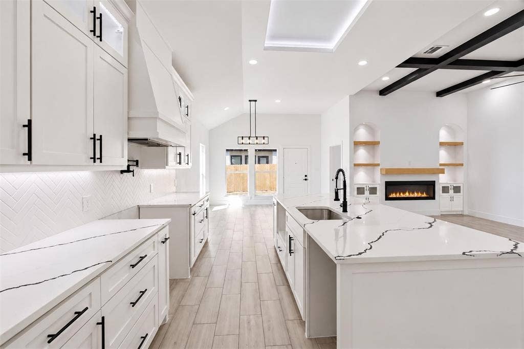 101 Connor Circle, Mabank, TX 75147 Kitchen featuring a waterfall-edge island with veined stone countertop, herringbone-pattern tile backsplash, wood-finish flooring, coffered ceiling detail, and built-in arched shelving with a fireplace