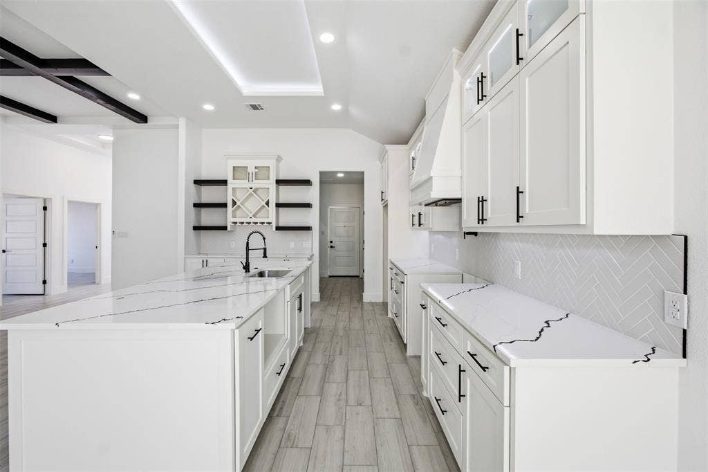 101 Connor Circle, Mabank, TX 75147 Kitchen featuring a large central island with veined stone-finish countertops, wood-finish flooring, white shaker-style cabinetry with matte black hardware, herringbone tile backsplash, and built-in wine storage