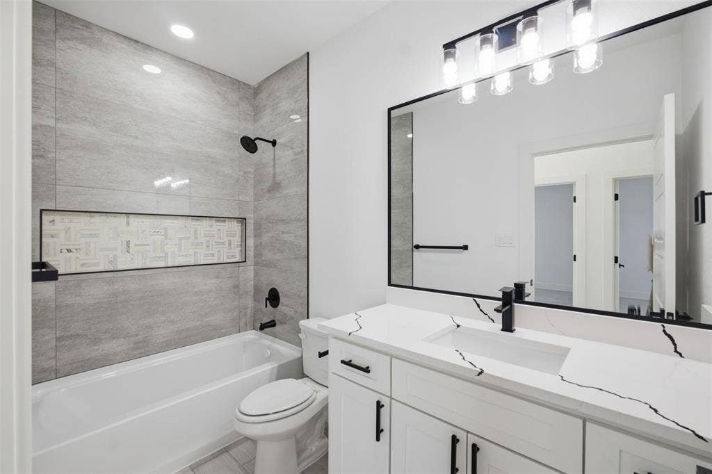 101 Connor Circle, Mabank, TX 75147 Bathroom featuring a white vanity with veined stone-finish countertop, undermount sink, matte black hardware, tiled tub surround with recessed decorative niche, and a large framed mirror with multi-bulb vanity lighting