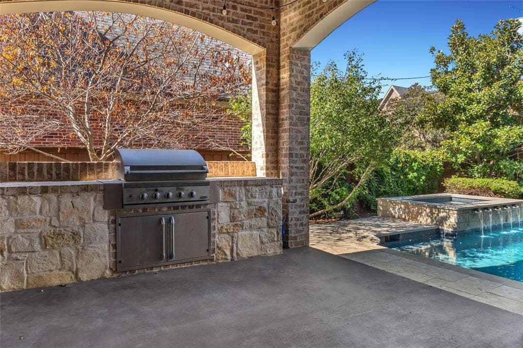 Covered patio featuring an arched brick ceiling, a built-in stone outdoor kitchen with stainless steel grill and storage, a flagstone pool deck, and an integrated spa with a water feature