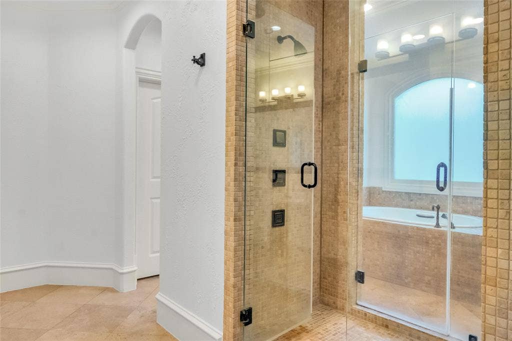 Walk-in shower featuring a frameless glass door, multi-function wall-mounted showerhead, and mosaic tile surround, paired with a deep soaking tub set beneath an arched window