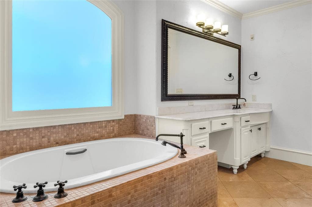 Soaking tub with mosaic tile surround, built-in vanity with stone countertop, oversized framed mirror, and arched window