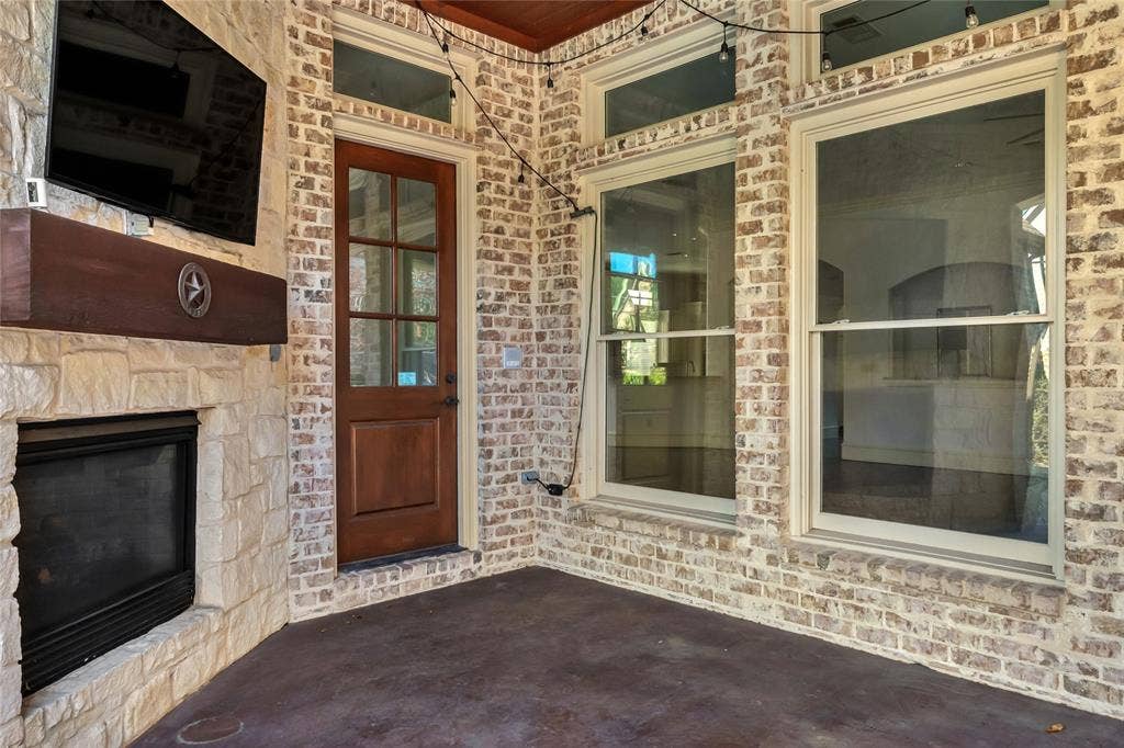 Covered patio featuring a stacked stone fireplace with a wood mantle, brick exterior walls, wood-finish door, and large rectangular windows with transoms