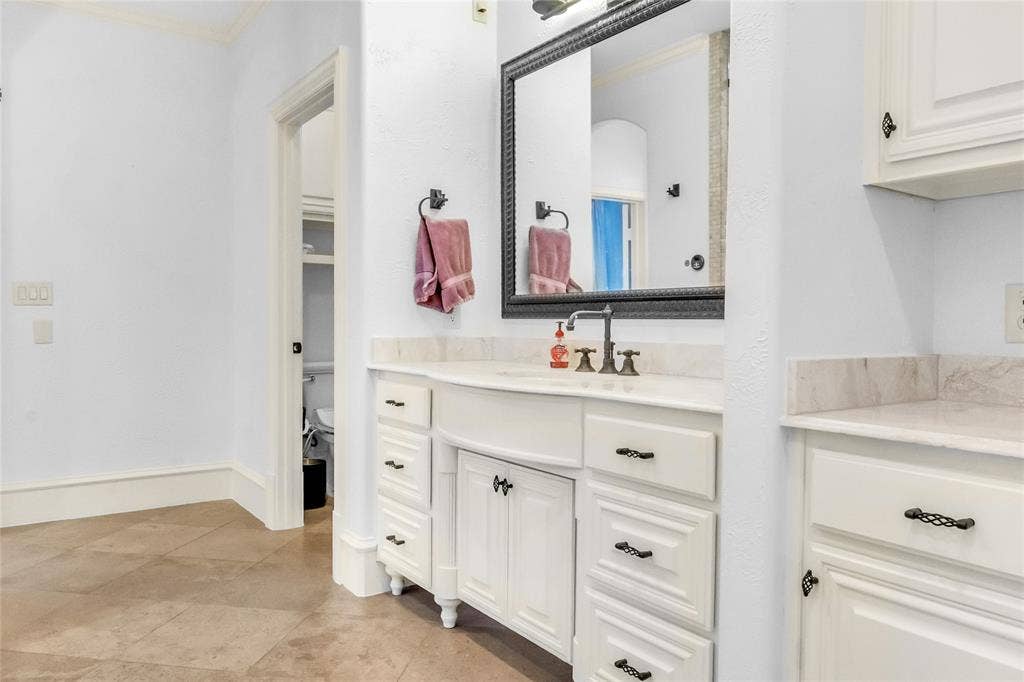 Custom vanity featuring white cabinetry, stone countertop with integrated sink, oil-rubbed bronze hardware, framed wall-mounted mirror, and large-format tile flooring