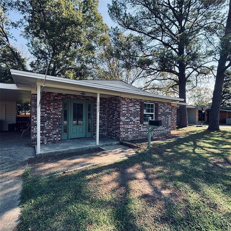 Single-story brick exterior featuring a covered front porch, attached carport, mature trees, and a front entry with glass-paneled sidelights