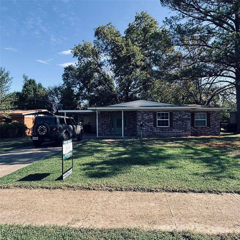 Single-story brick exterior featuring a covered carport, concrete driveway, and mature trees