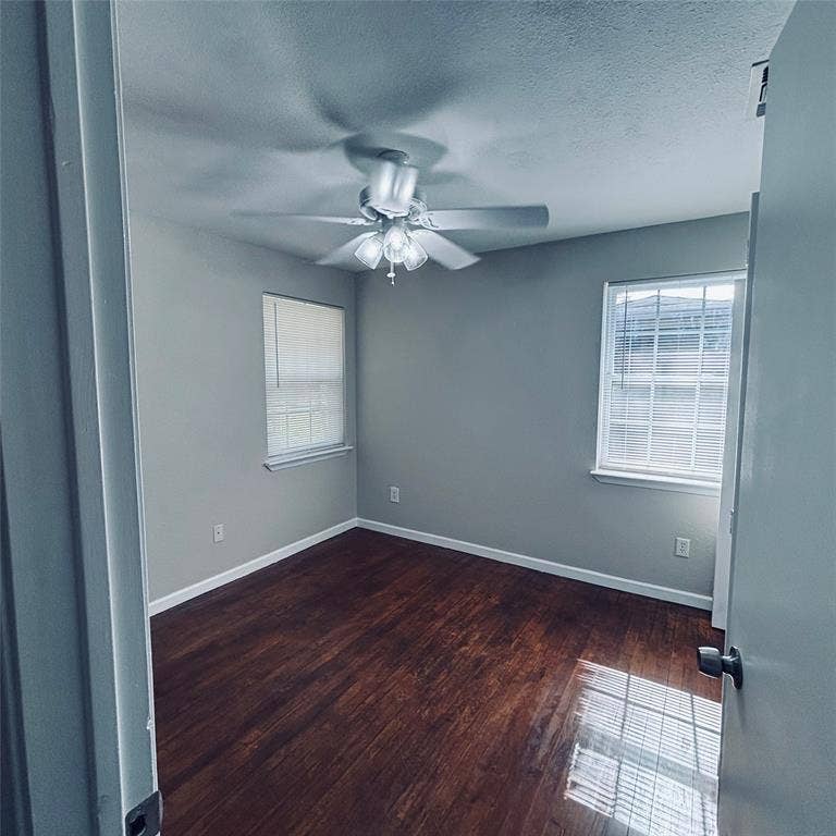Room featuring dark wood-finish flooring, two windows with horizontal blinds, and a central ceiling fan with integrated lighting