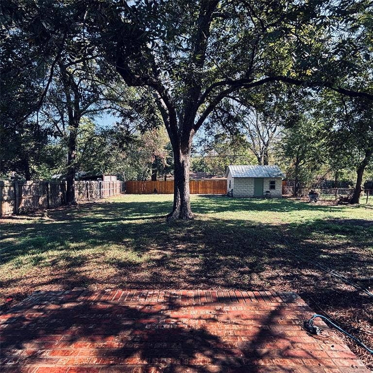 Rear grounds featuring a brick patio, mature canopy trees, a detached storage structure with a metal roof, and perimeter wood fencing