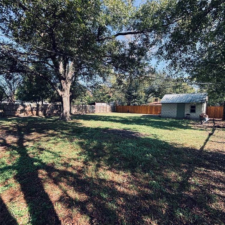 Expansive backyard featuring mature shade trees, a stone-exterior storage building with a metal roof, and perimeter wood fencing