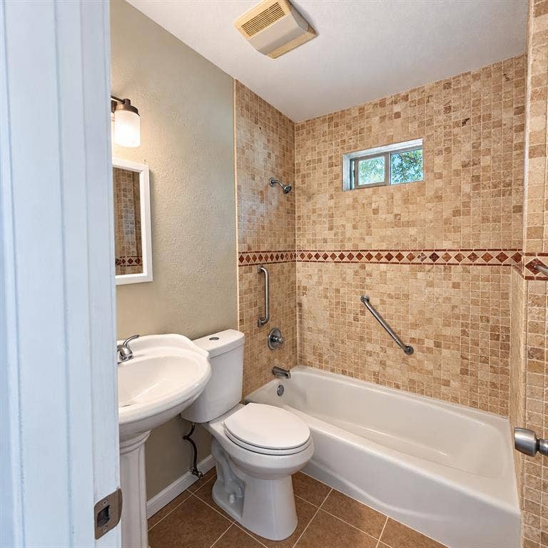 Bathroom featuring a pedestal sink, porcelain tub, mosaic tile surround with a decorative border, and wall-mounted metal grab bars