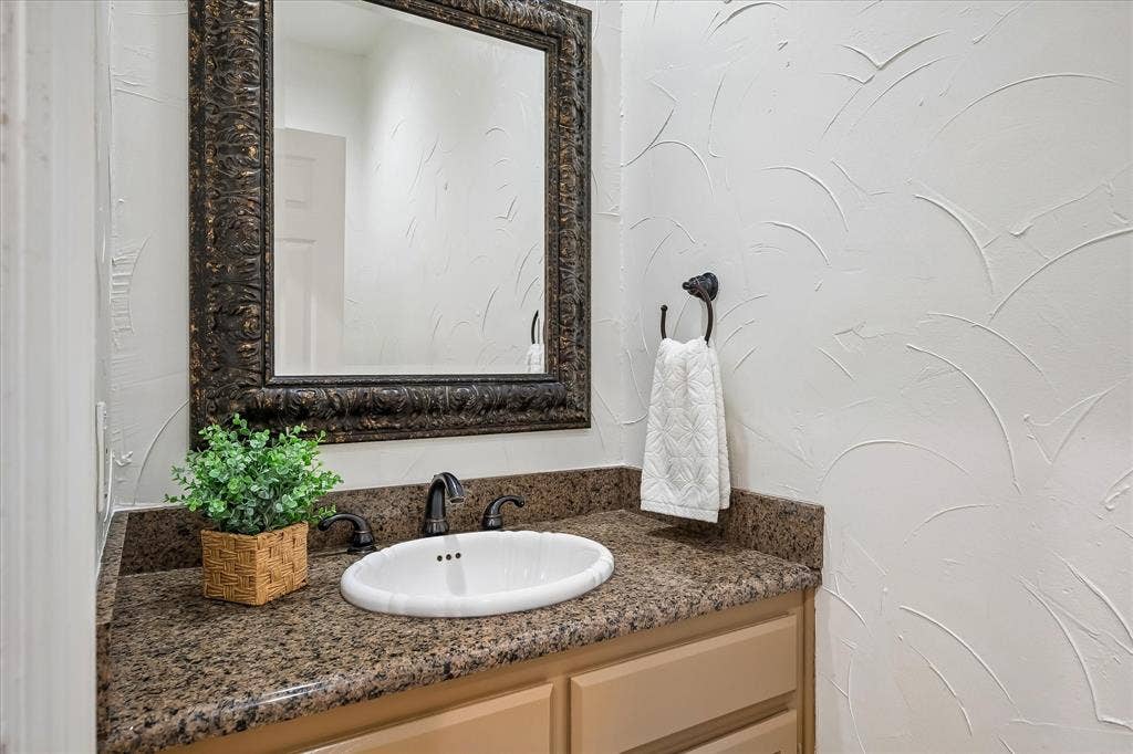 4611 O Connor Court, Irving, TX 75062 Vanity featuring stone-finish countertop, oval drop-in sink, dark metal widespread faucet, decorative framed mirror, and textured wall finish