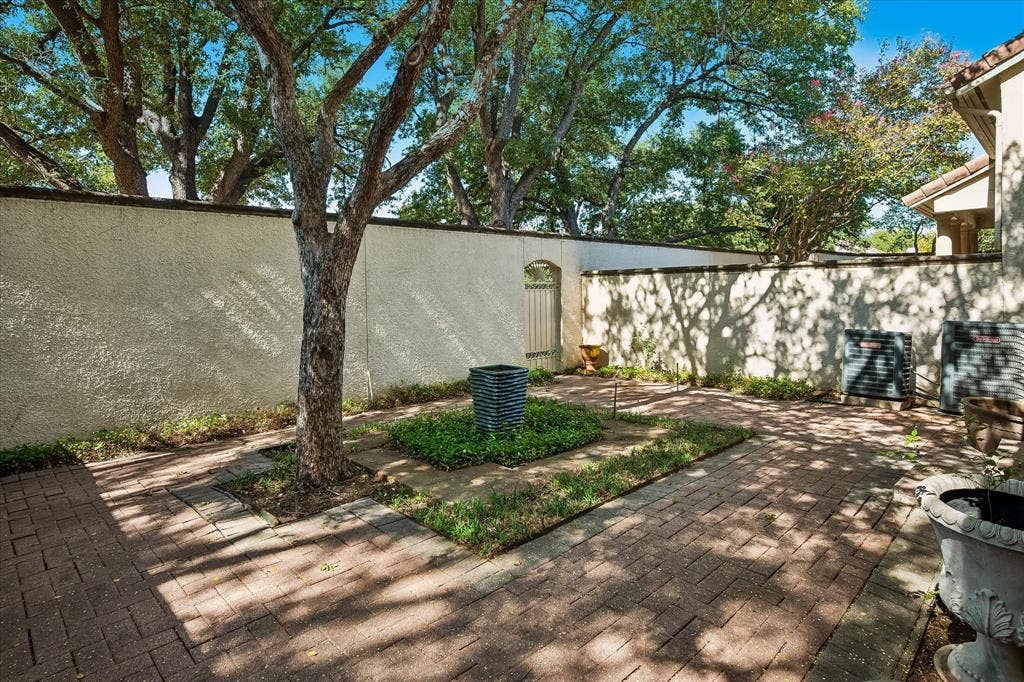 4611 O Connor Court, Irving, TX 75062 Enclosed courtyard featuring decorative brick pavers, mature trees, a central ground-level planter, and a perimeter stucco wall with a metal gate