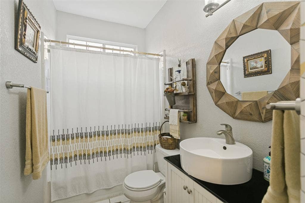 3212 Chippenham Drive, Plano, TX 75093 White oval vessel sink with a brushed-finish faucet, dark countertop, white beadboard vanity cabinet, and brushed-finish wall-mounted light fixture