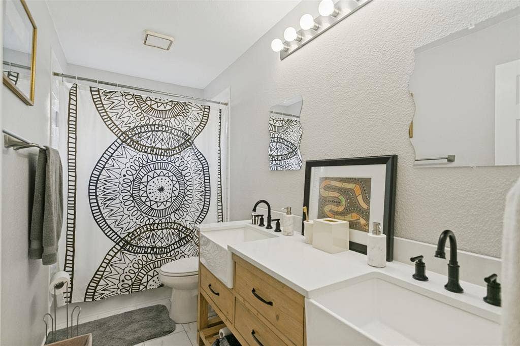 3212 Chippenham Drive, Plano, TX 75093 Bathroom featuring a custom wood-finish vanity with dual farmhouse sinks, light-toned countertop, matte black fixtures, and a multi-bulb vanity light