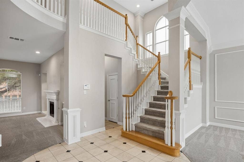Foyer featuring tile flooring with decorative inserts, a multi-story staircase with a wooden handrail and white balusters, an arched window, crown molding, and a fireplace with a white mantel