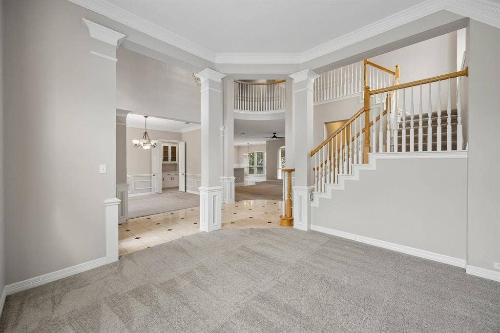 Double-height foyer featuring decorative columns, an open staircase with a wooden handrail, crown molding, and tiled flooring with inset accents