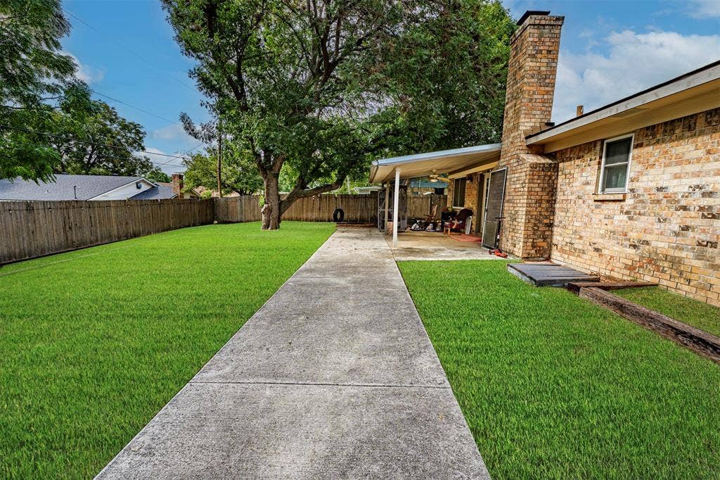 Backyard featuring a brick masonry exterior, a covered concrete patio, a central concrete walkway, a wood privacy fence, and mature trees