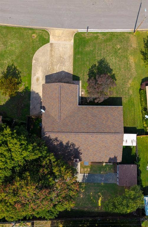 Aerial view featuring an asphalt shingle roof, concrete driveway, expansive grass lawn, and mature trees on a corner lot