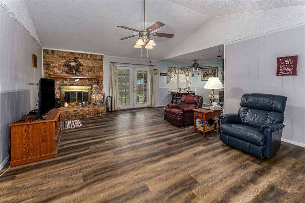 Vaulted ceiling featuring a central lighted ceiling fan, wood-finish flooring, masonry fireplace with a wood mantel, and glass-paned double doors