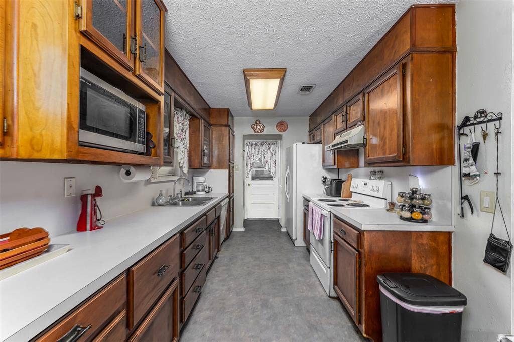 Galley kitchen featuring wood cabinetry, solid-surface countertops, dual-basin stainless steel sink, and integrated overhead lighting