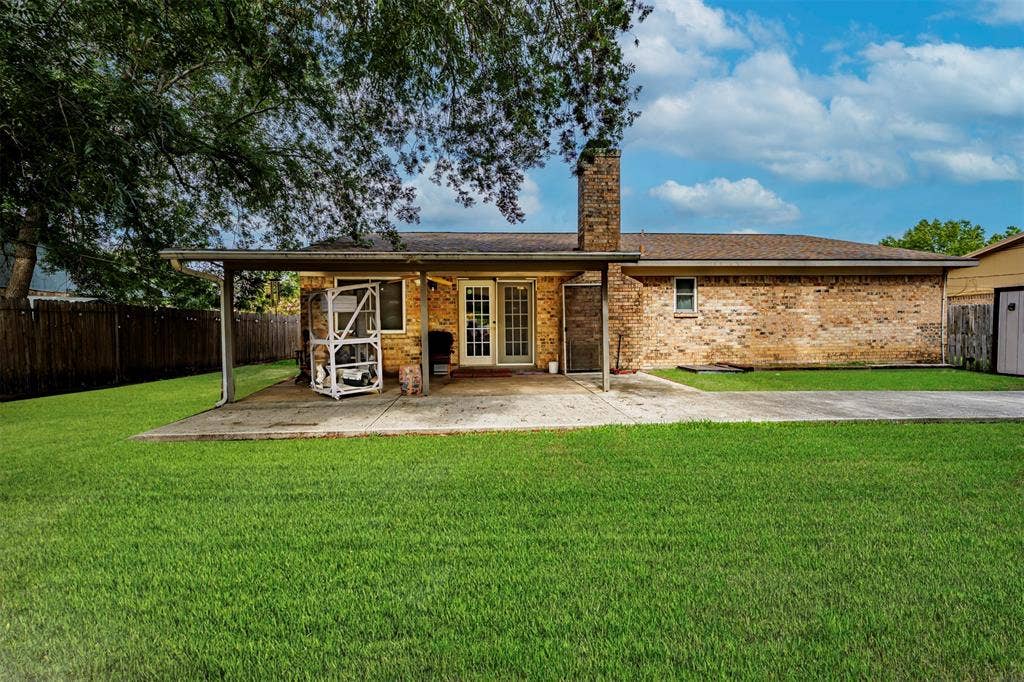 Brick exterior featuring a covered concrete patio, an expansive lawn, a stone chimney, and wood-panel fencing