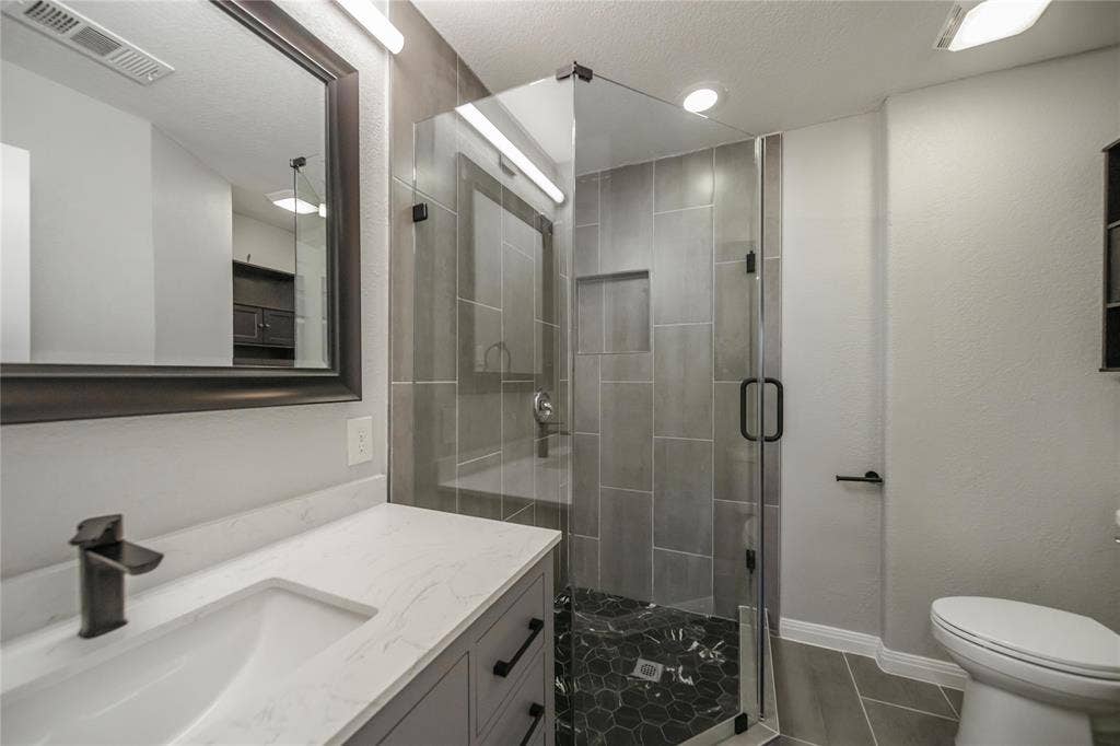 Bathroom featuring a vanity with a white countertop and rectangular undermount sink, matte black faucet, glass-enclosed shower with tiled walls and a built-in recessed niche, and dark geometric floor tile