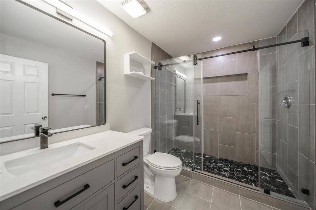 Bathroom featuring a vanity with light-toned countertop, rectangular undermount sink, and matte black hardware, paired with a glass-enclosed shower, oversized tile walls, and a patterned hexagonal tile shower floor
