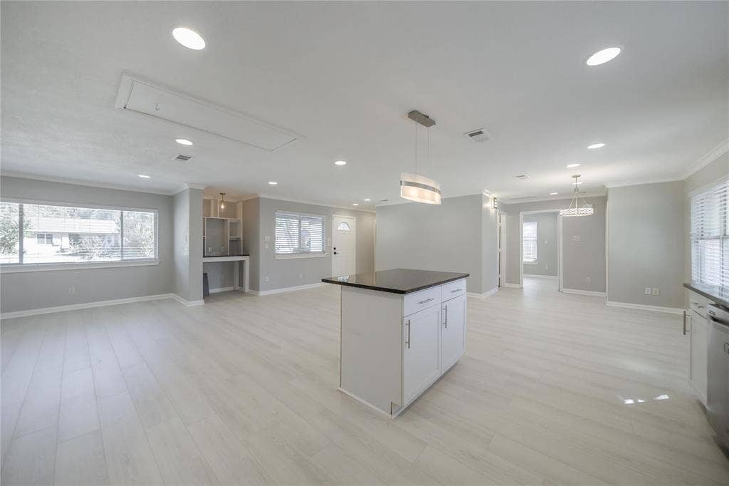 Open-concept interior featuring light wood-finish flooring, recessed lighting, a central kitchen island with dark stone countertop, and two tiered pendant light fixtures