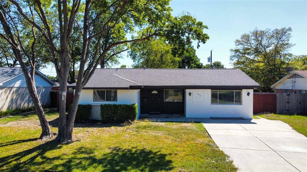 Single-story residence featuring a painted masonry exterior, composite shingle roof, concrete driveway, and mature landscaping