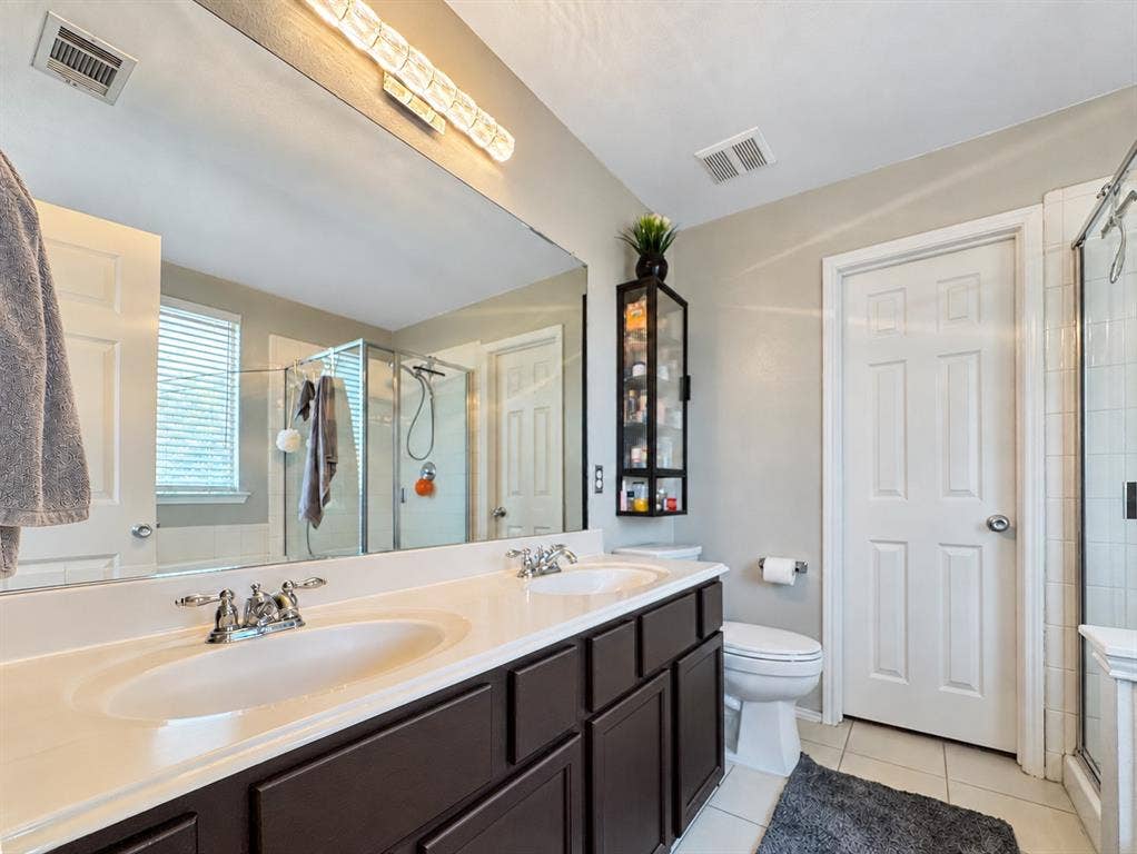 Double vanity featuring a light-toned countertop, dark-finished cabinetry, a full-width mirror, integrated lighting, and a glass-enclosed shower