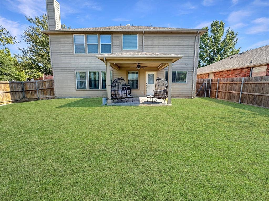 Rear elevation featuring a covered patio with a ceiling fan, horizontal lap siding, and an expansive grass lawn enclosed by a wood privacy fence