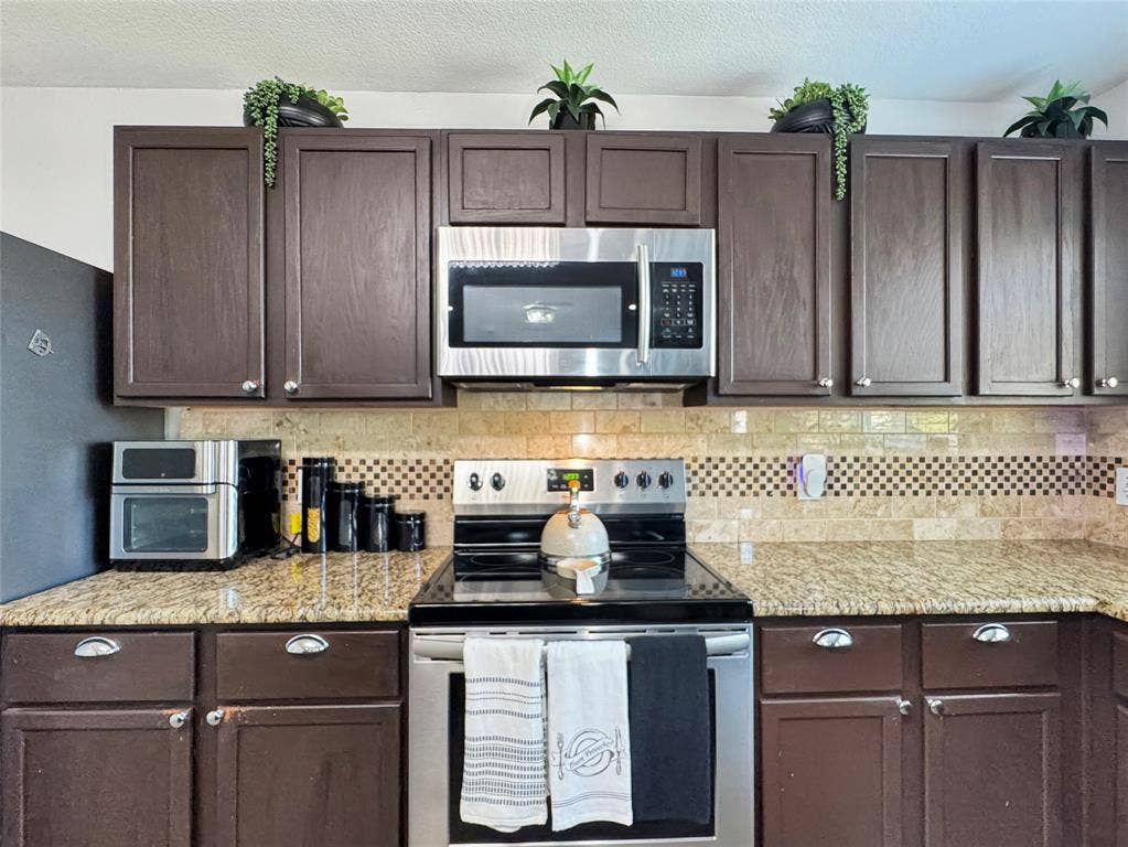 Dark wood cabinetry, stone countertops, stone tile backsplash with decorative mosaic accent, and stainless steel kitchen appliances