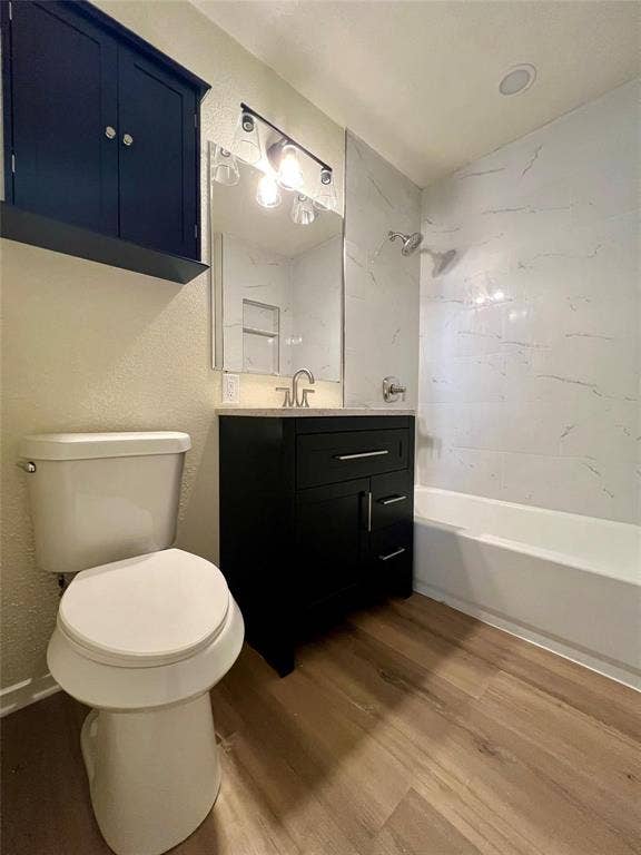 Bathroom featuring a dark wood-finish vanity, light-toned stone-finish countertop, wall-mounted storage cabinetry, wood-finish flooring, and a tub enclosure with light-toned wall tile