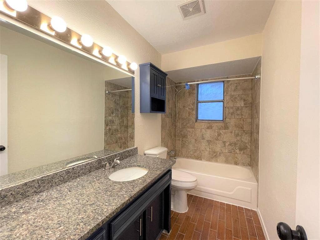 Bathroom featuring a vanity with speckled stone-finish countertop and oval undermount sink, dark cabinetry, wood-finish tile flooring, and tub surround with decorative wall tile