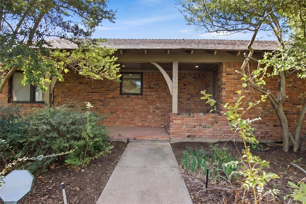 7606 Currin Drive, Dallas, TX 75230 Single-story exterior featuring a brick facade, covered porch, arched support brackets, and concrete walkway