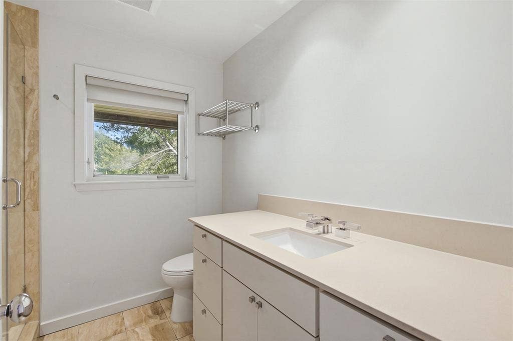 7606 Currin Drive, Dallas, TX 75230 Bathroom featuring a light-toned vanity with an integrated countertop and sink, stone-tile flooring, a glass-enclosed shower with stone surround, and a wall-mounted metal shelving unit