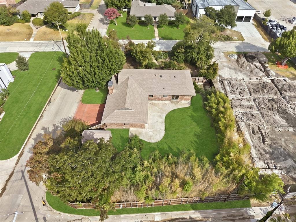 7606 Currin Drive, Dallas, TX 75230 Aerial perspective of a single-story brick residence featuring a paved driveway, a multi-section roof, a concrete rear patio, and established mature trees