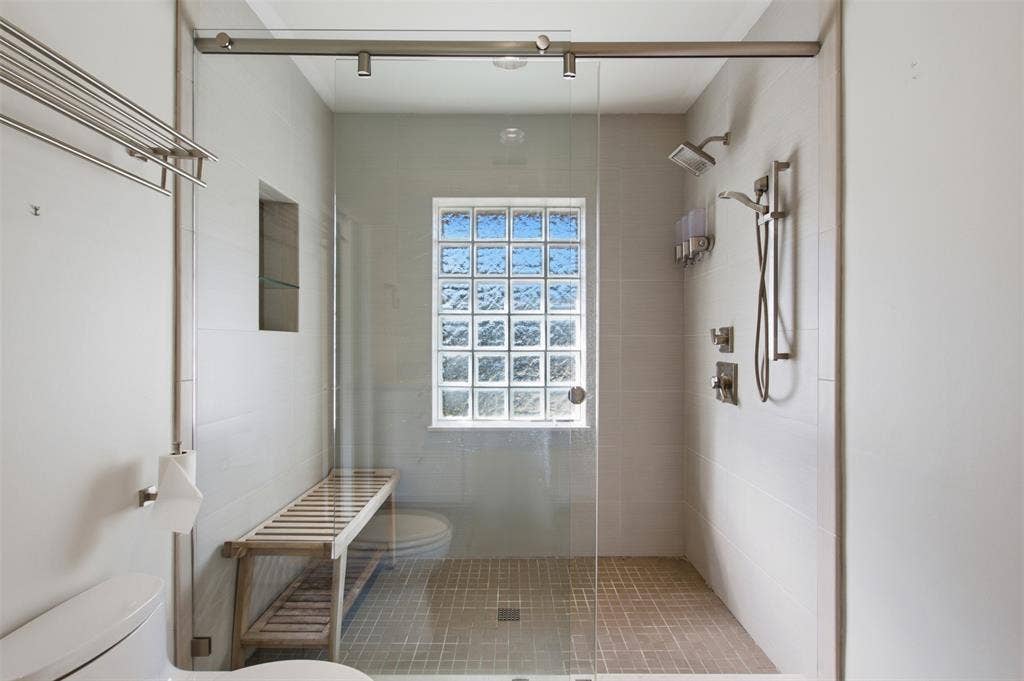 7606 Currin Drive, Dallas, TX 75230 Walk-in shower featuring a glass enclosure with a sliding door, dual shower heads, a built-in recessed niche, and a glass block window
