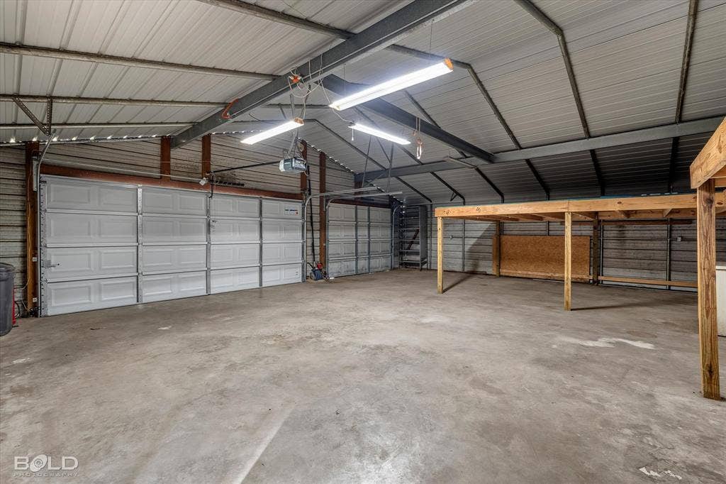 Spacious metal-frame structure featuring concrete flooring, multiple overhead garage doors, high-output fluorescent lighting, and elevated wooden platform storage