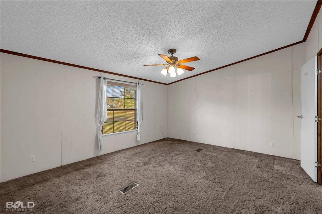 Interior space featuring textured ceilings, dark carpeting, wood-finish crown molding, a central ceiling fan with integrated lighting, and a single-pane window