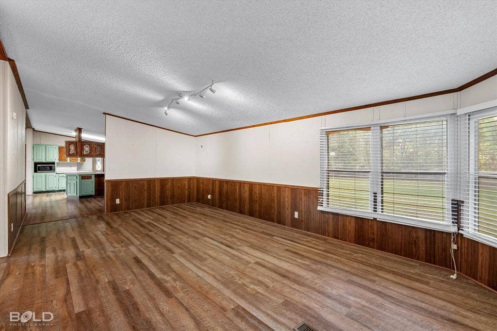 Living area featuring wood-finish flooring, wood panel wainscoting, ceiling-mounted track lighting, and multiple windows with horizontal blinds