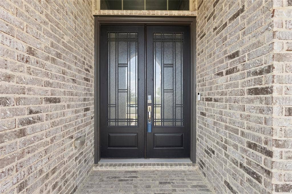 Double entry doors featuring dark-toned frames, geometric glass inserts, and a transom window, surrounded by neutral-toned brick masonry