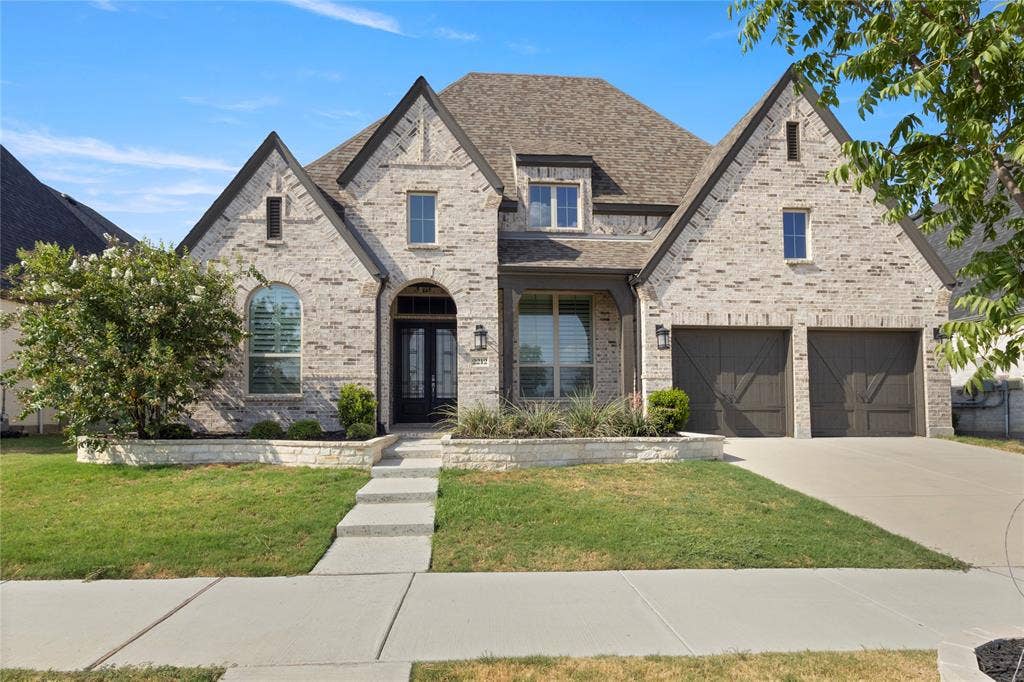 Traditional brick facade featuring multiple roof gables, arched entry, stone-accented landscape borders, and double-bay garage with decorative carriage-style doors