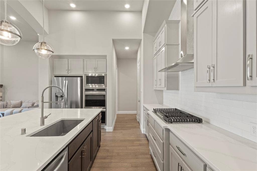 Kitchen featuring a spacious island with a single-basin undermount sink, quartz countertops, light-toned cabinetry, stainless steel built-in appliances, and wood-finish flooring